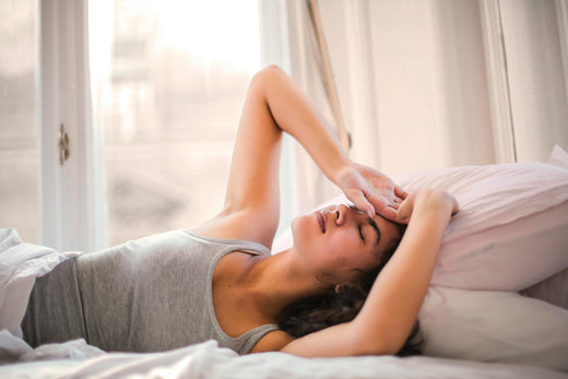 A woman wearing a grey sleeping vest laying in bed, half covered by the sheets with her eyes closed.